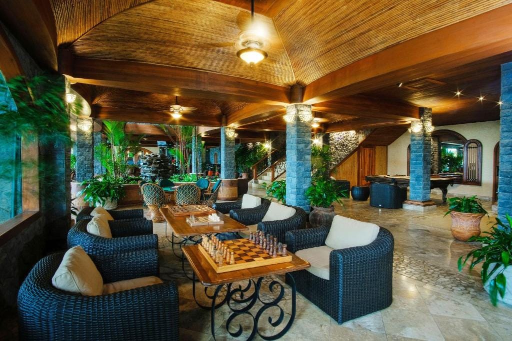 Spacious, elegant lounge with wicker chairs, a chessboard on a wooden table, lush green plants, stone columns, a billiard table in the background, and a wooden ceiling with ceiling fans and warm lighting.
