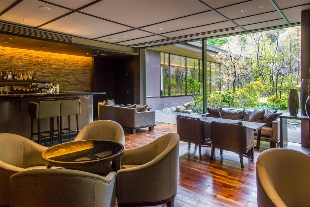 Modern lounge with beige armchairs, sofas, and a small bar. Large windows reveal a green, landscaped garden outside. The space features wooden floors and a warm, inviting atmosphere.