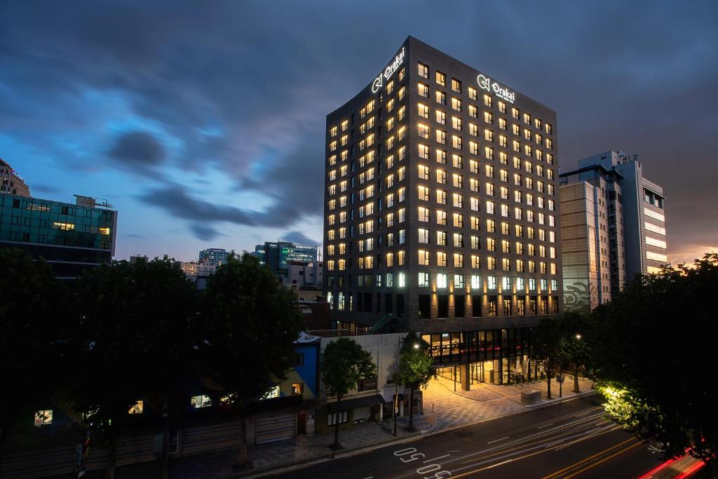A tall, modern hotel building with many illuminated windows stands beside a busy city street at dusk, with surrounding trees and nearby office buildings under a dramatic, cloudy sky.