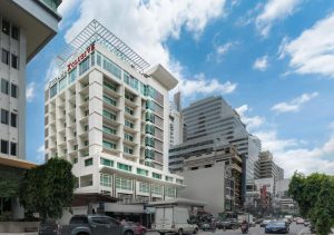 Modern multi-story hotel building with balconies, labeled “Red Planet,” stands on a busy street lined with cars, motorbikes, and other tall office buildings under a partly cloudy sky.