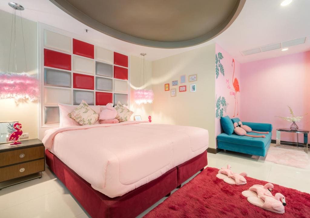 A brightly lit bedroom with a pink and red color scheme, featuring a large bed with pink bedding, flamingo decor, plush toys, a blue sofa, and wall art. The room has playful and cozy elements throughout.