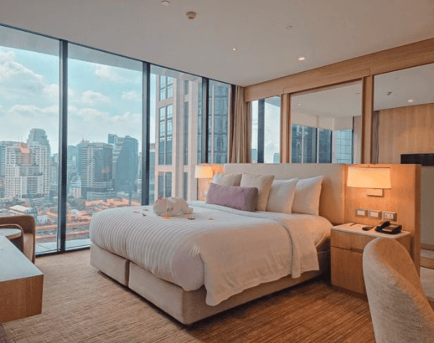 A modern hotel room with a large bed, side tables with lamps, a desk, and floor-to-ceiling windows offering a city skyline view during the day.