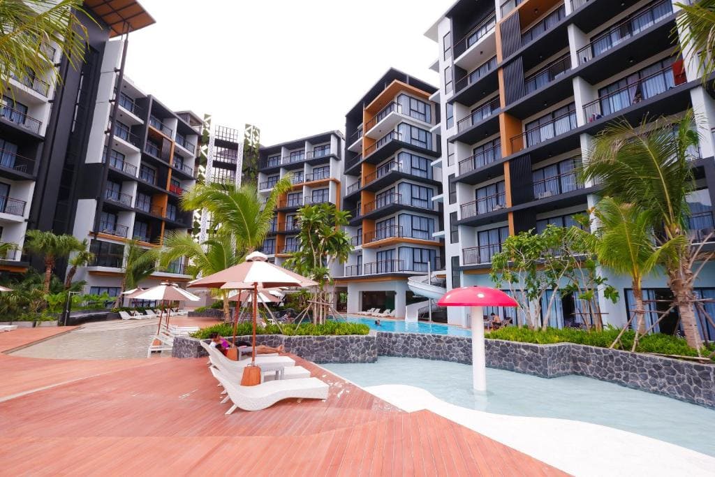Modern multi-story resort buildings surround a pool area with lounge chairs, umbrellas, palm trees, and a red-and-white mushroom fountain. The deck is wooden and the atmosphere is tropical and inviting.