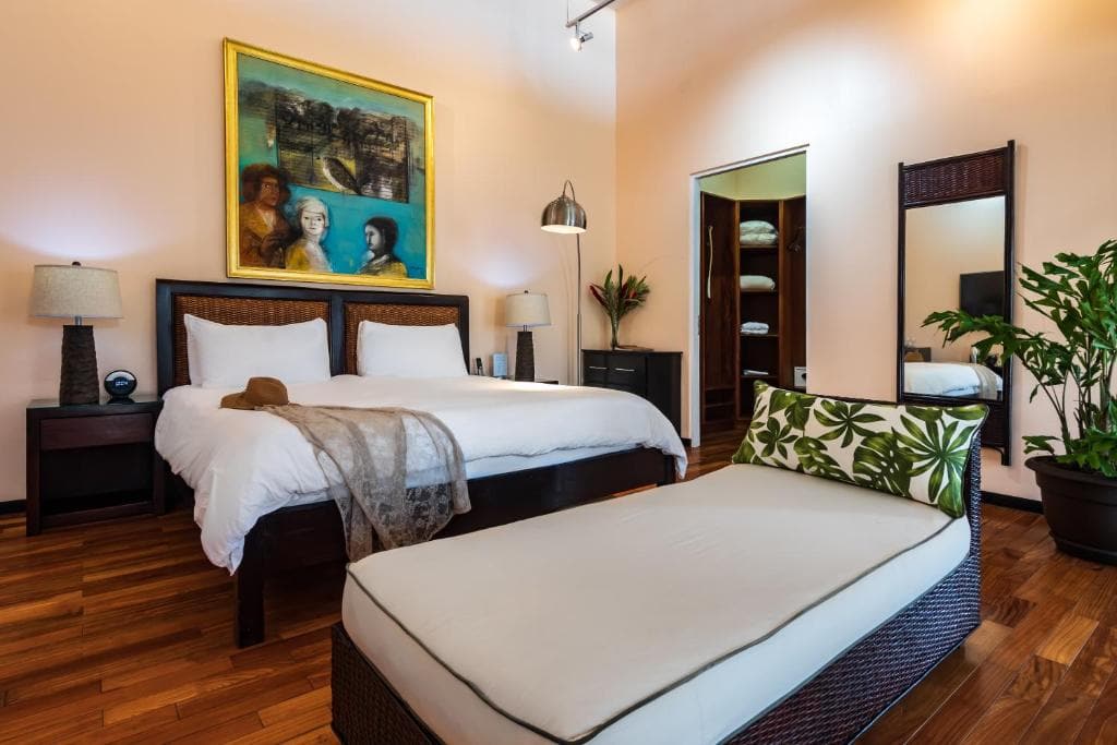 A cozy bedroom with a large bed, white bedding, a lounge chair with a leaf-patterned pillow, wooden floors, a potted plant, a tall mirror, and a colorful painting hanging above the bed.