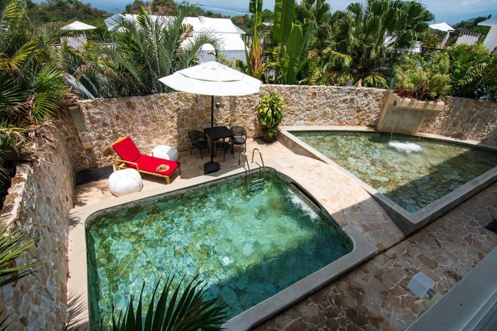 A tropical outdoor area with two small clear pools, surrounded by stone walls and lush green plants. A red lounge chair, two white beanbags, a black table, and a white umbrella are next to the pools.