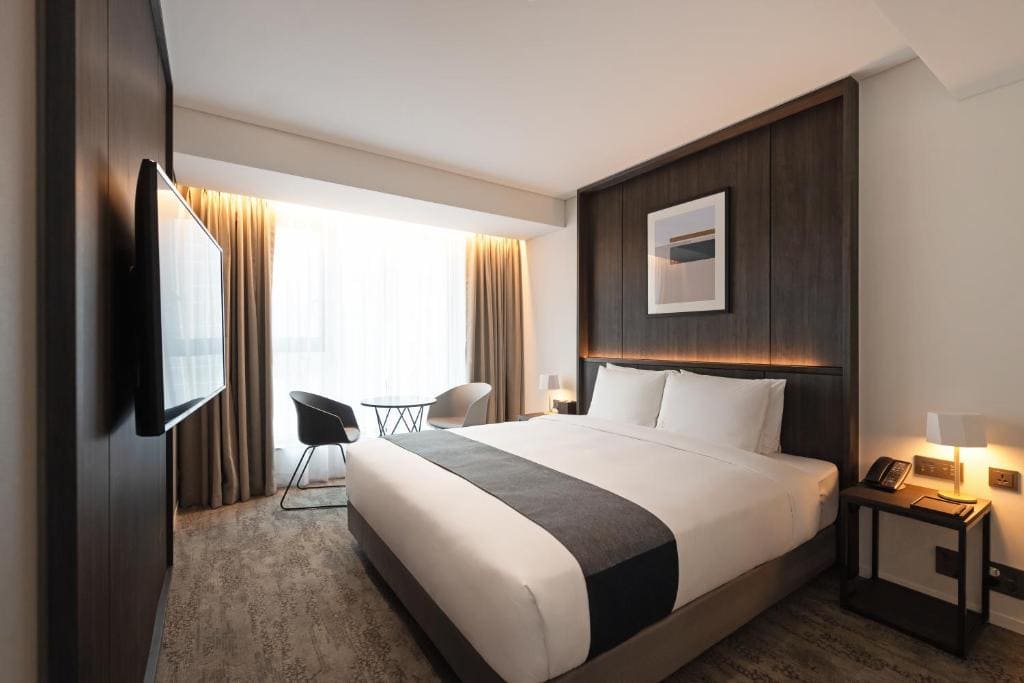 Modern hotel room with a large bed, dark wood accents, a wall-mounted TV, a small round table with two chairs by a window, and bedside tables with lamps and a phone. Soft natural light fills the space.