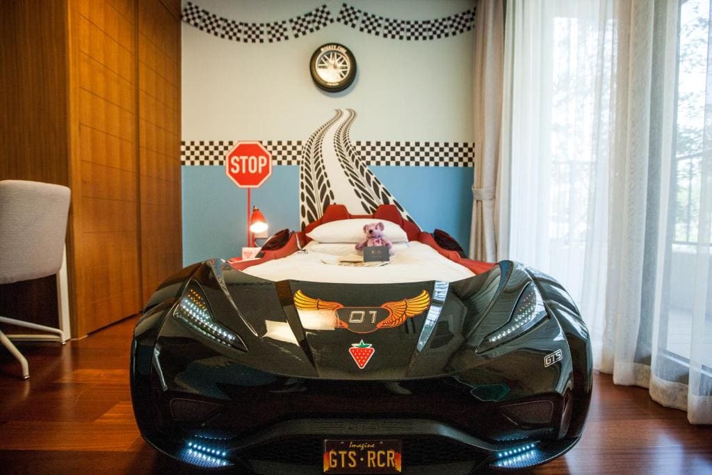 A children’s bedroom features a car-shaped bed with racing decals, a checkered wall design, a stop sign, a desk and chair, and a window with sheer curtains. A stuffed animal sits on the bed.