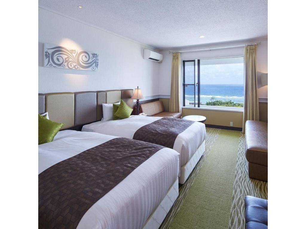 Hotel room with two double beds, green accent pillows, a lamp, and a window with sheer curtains offering a view of the ocean and blue sky. There is also a small table, chair, and sofa.