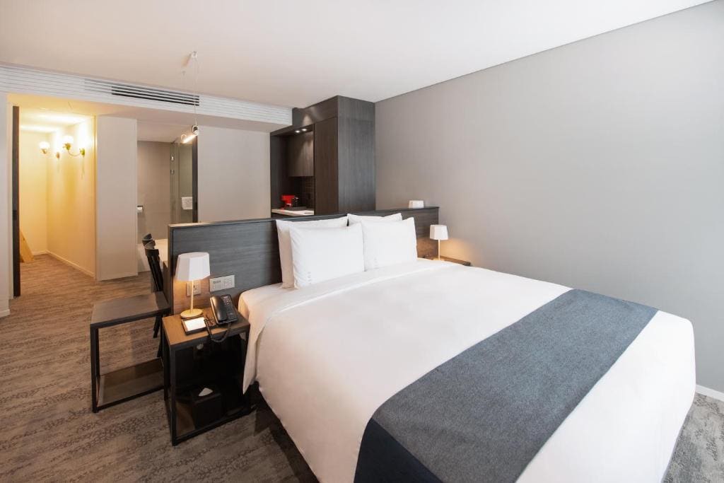 A modern hotel room with a large neatly made bed, white linens, dark wood headboard, bedside tables with lamps, a desk, and a hallway leading to the entrance. The decor is minimalist and contemporary.