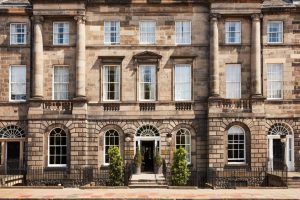 A symmetrical stone building with tall columns, large sash windows, arched doorways, and two small trees flanking the main entrance, viewed from the street.