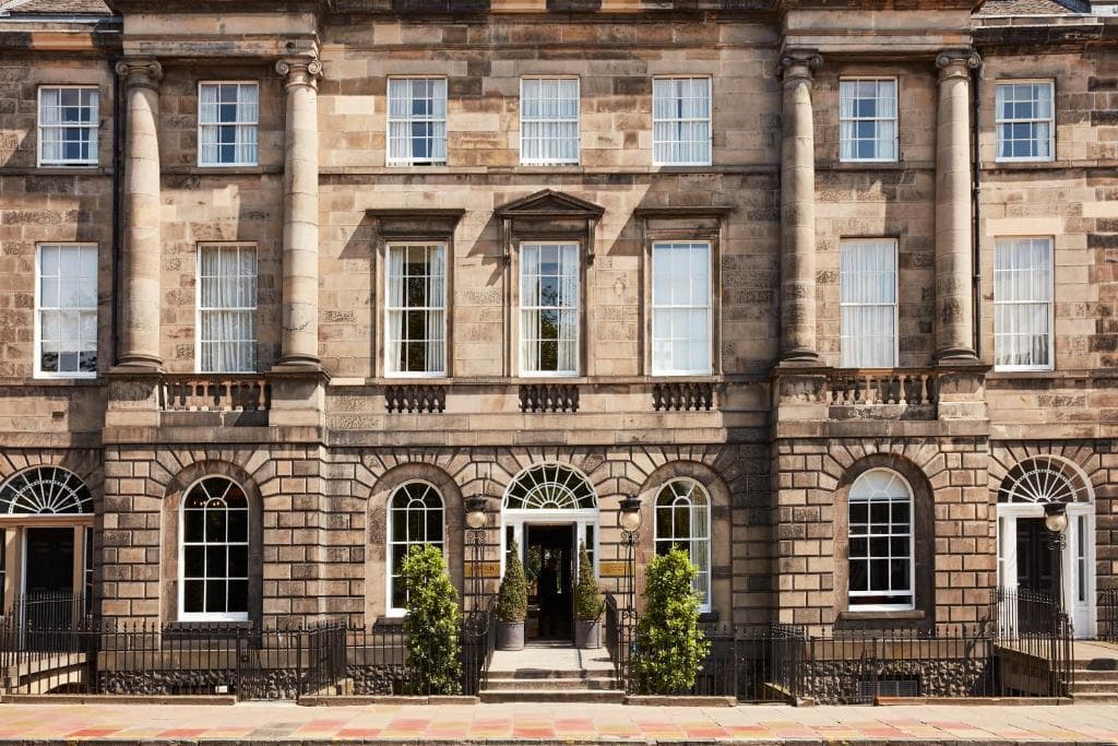 A symmetrical stone building with tall columns, large sash windows, arched doorways, and two small trees flanking the main entrance, viewed from the street.