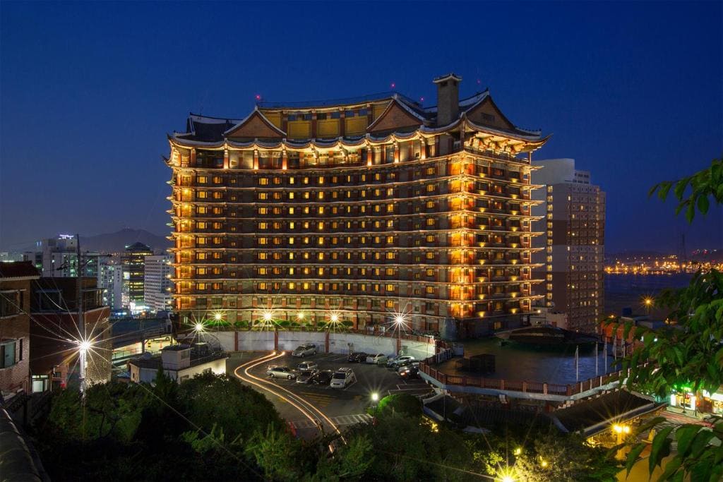 A large, ornate hotel with traditional Asian architectural features is brightly illuminated at night, standing against a dark blue sky in a cityscape with other buildings and distant lights.