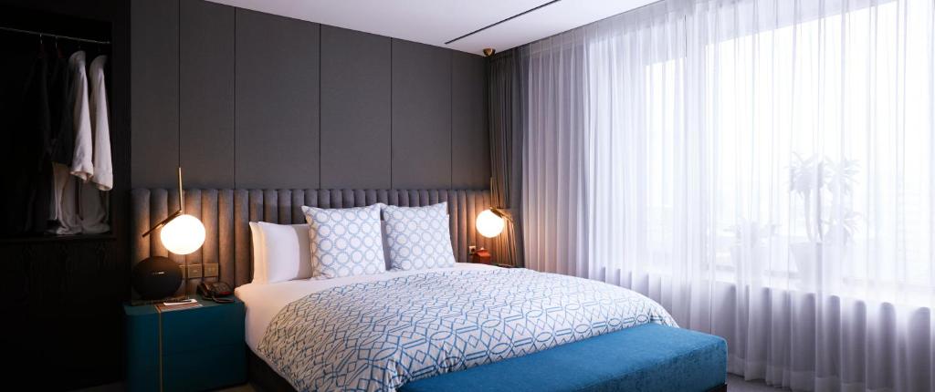 Modern bedroom with a double bed, blue geometric bedding, two round bedside lamps, a teal nightstand, floor-to-ceiling curtains, and a large window letting in natural light.