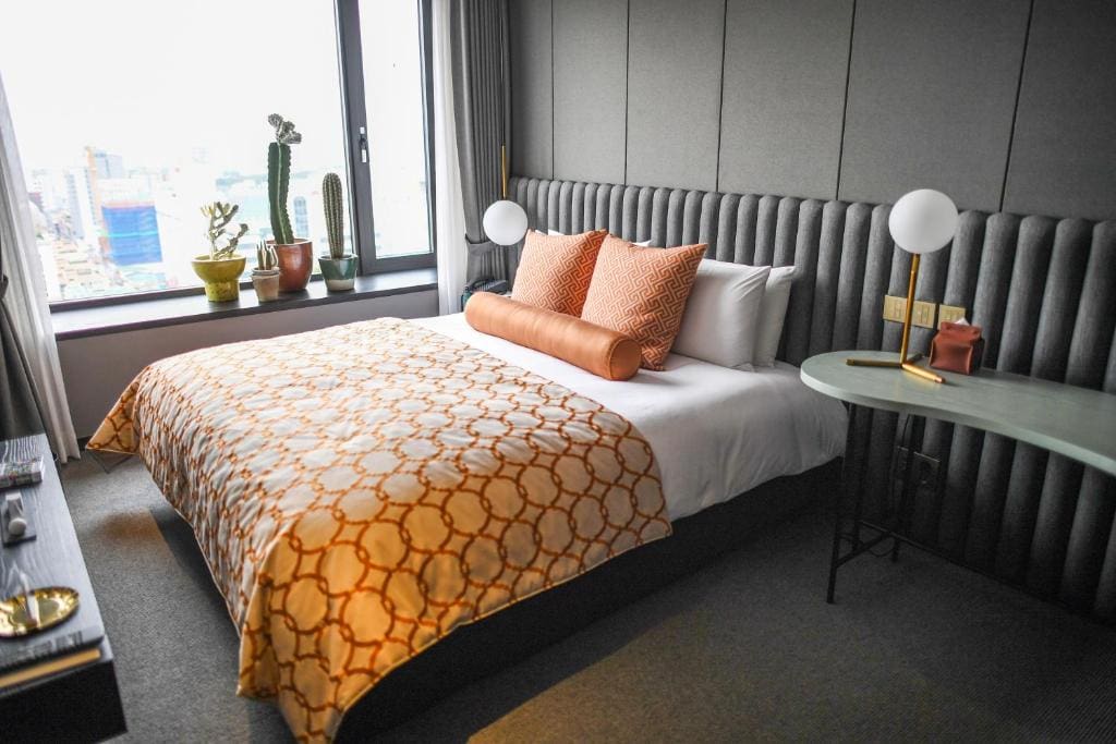 A modern hotel room with a neatly made bed featuring orange-patterned bedding, orange pillows, a bedside table with a round lamp, and a window with potted cacti and a city view.