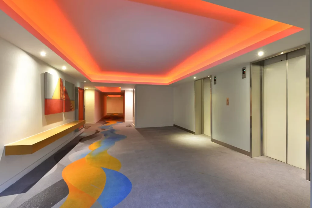A modern hallway with two elevators, colorful abstract carpet, a wall-mounted artwork, and a recessed ceiling with orange lighting. The walls are white, and the space is brightly lit.