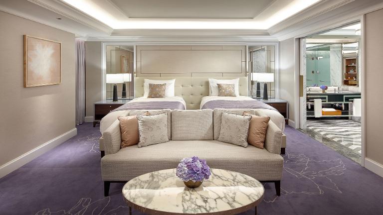Luxurious hotel room with two beds, a cushioned bench, and a marble coffee table with a vase of purple flowers. The room features soft lighting, modern decor, and an open view of a spacious bathroom.