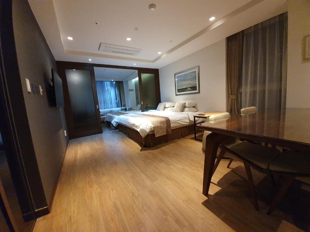 A modern hotel room with two double beds, a wooden floor, a table with chairs, large windows with sheer curtains, and a painting on the wall above the beds. The room is well-lit and spacious.