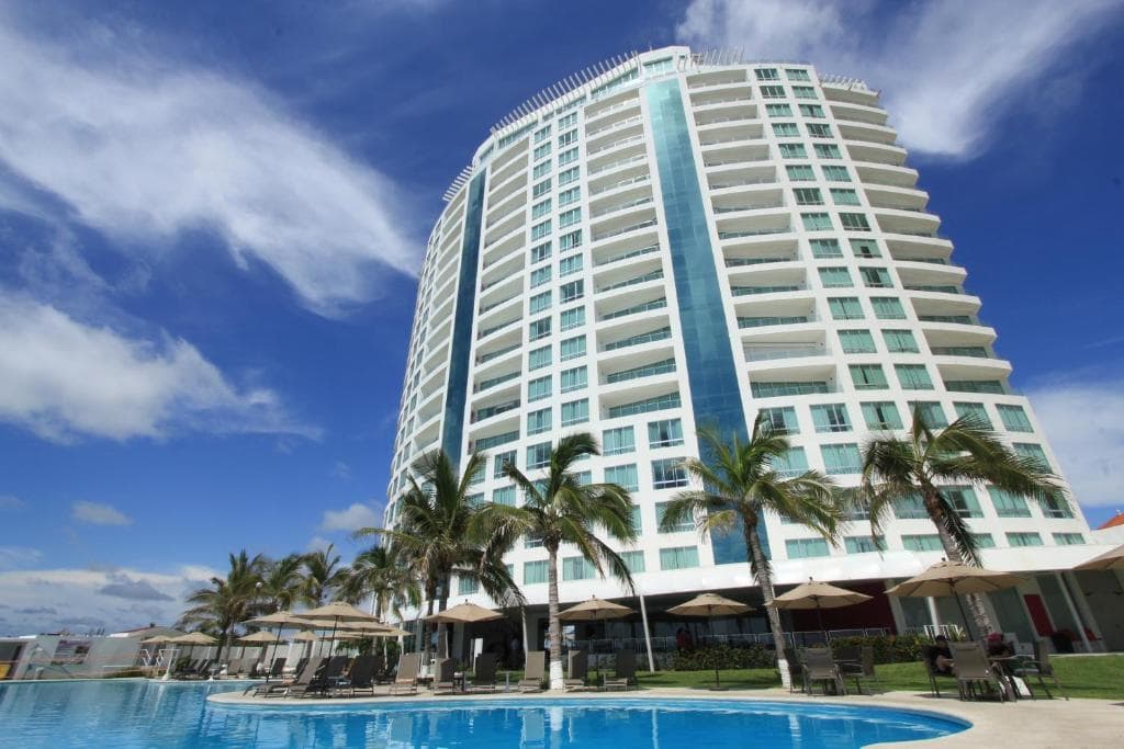 A tall white modern hotel with balconies sits beside a clear blue swimming pool, surrounded by palm trees and lounge chairs under umbrellas, under a bright blue sky with scattered clouds.