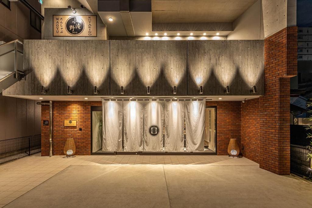 A modern restaurant exterior with brick and concrete walls, warm overhead lighting, and white curtains hanging at the entrance. A circular logo is displayed on the curtains, and two decorative lamps sit on either side of the doorway.
