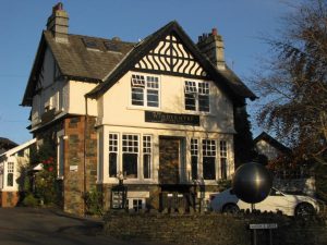 A two-story, cream-colored guesthouse with timber accents and a sign reading Windermere above the door. A white car is parked outside, and bare trees and a round metal sculpture are visible near a stone wall.