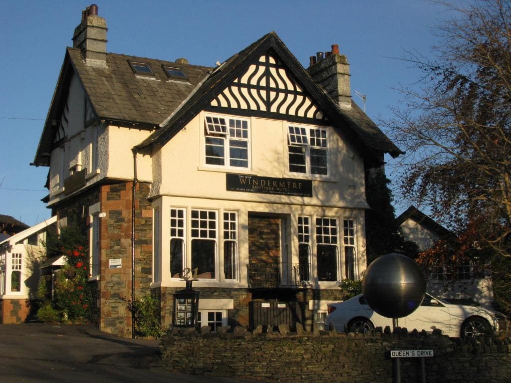 A two-story, cream-colored guesthouse with timber accents and a sign reading Windermere above the door. A white car is parked outside, and bare trees and a round metal sculpture are visible near a stone wall.