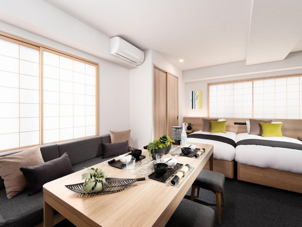 A modern, minimalistic hotel room with two beds, a wooden dining table set for four, a gray sofa with cushions, decorative plants, and large windows with shoji screens, creating a bright and inviting atmosphere.