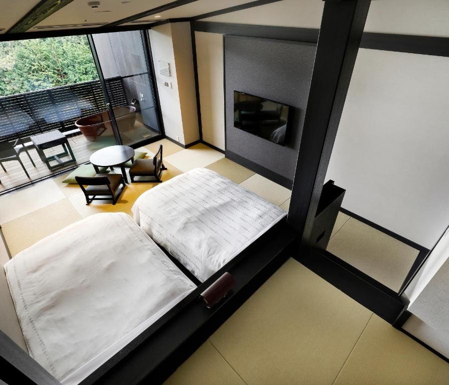 A modern Japanese-style hotel room with two beds on tatami mats, a round table with two chairs, a flat-screen TV, large windows, and a balcony with outdoor seating and a view of greenery.