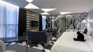 Modern hotel room with bold black-and-white geometric patterns on walls, floor, and ceiling. Two beds with white linens, a black cat on one bed, a seating area with dark sofas, and large windows with sheer curtains.