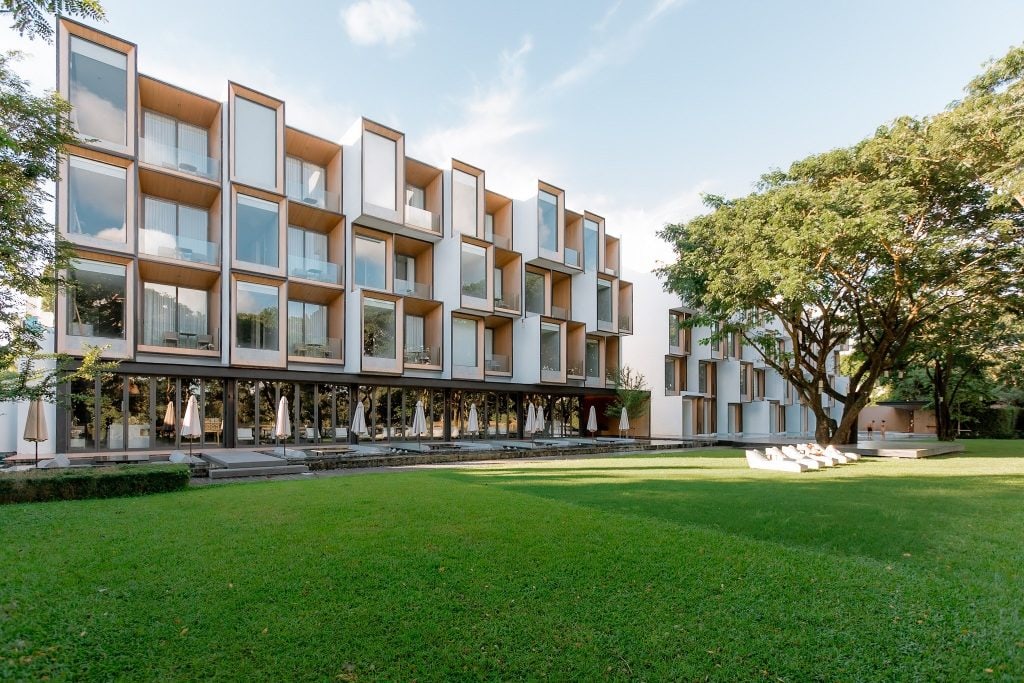 A modern four-story building with large glass windows and box-like balconies overlooks a spacious green lawn with trees and outdoor seating under umbrellas. The sky is clear with a few clouds.