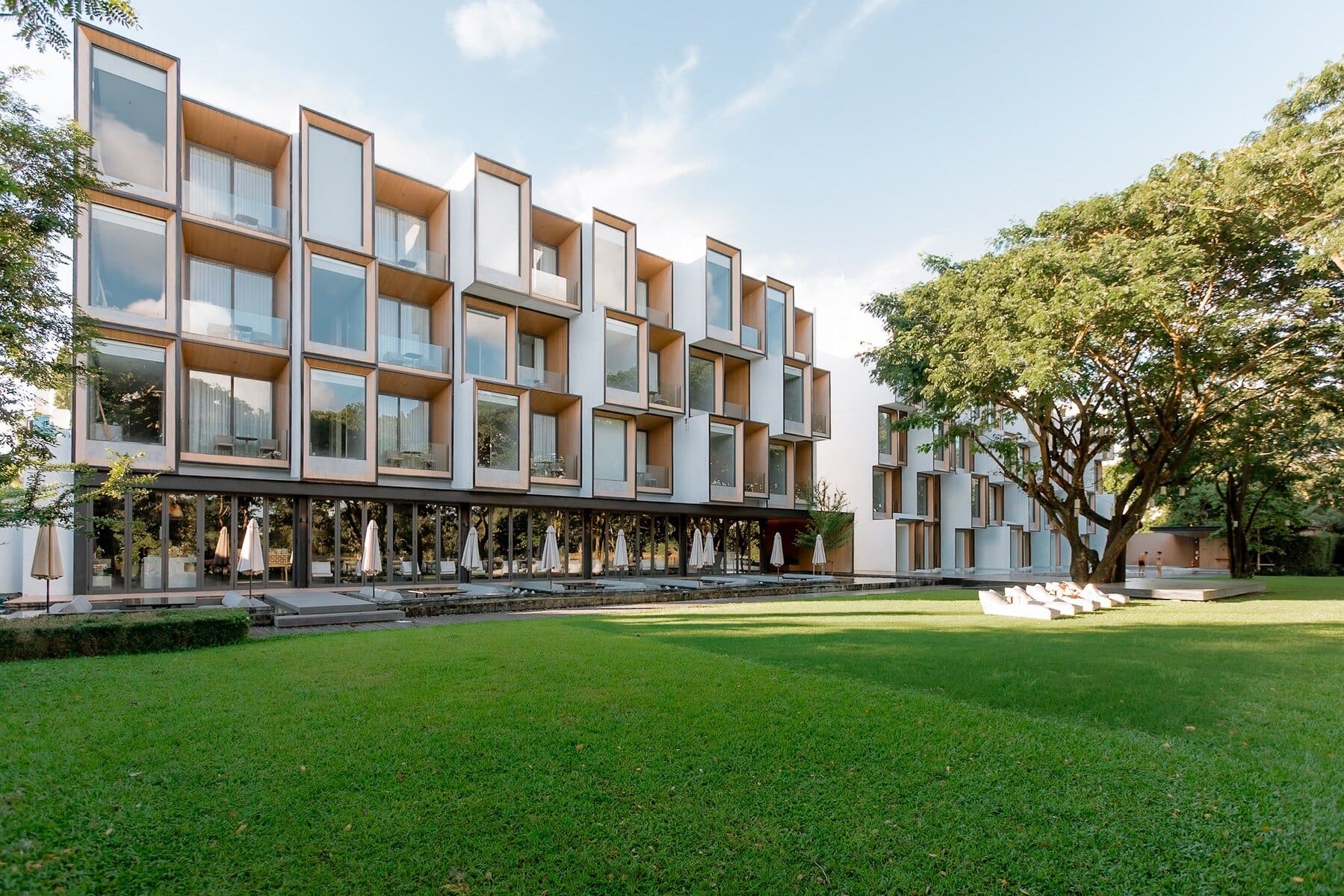A modern four-story building with large glass windows and box-like balconies overlooks a spacious green lawn with trees and outdoor seating under umbrellas. The sky is clear with a few clouds.
