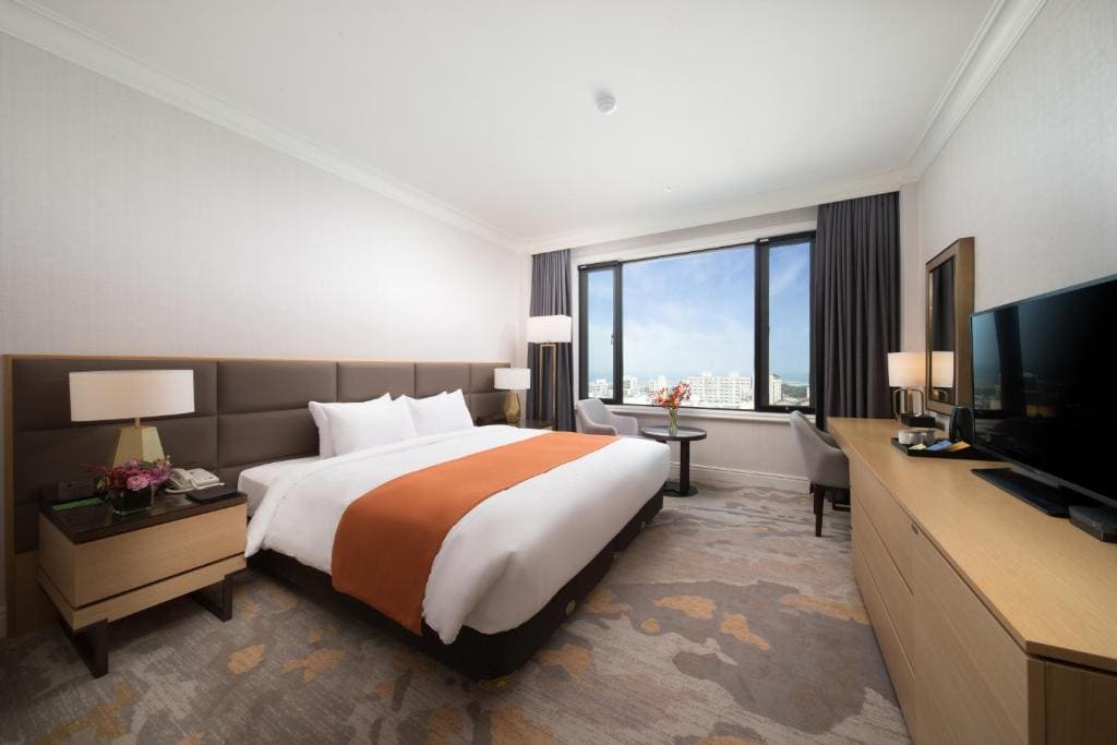 Modern hotel room with a large bed, orange throw blanket, bedside tables with lamps, a TV on a wooden dresser, two chairs with a small table by a window showing a city view, and neutral decor.