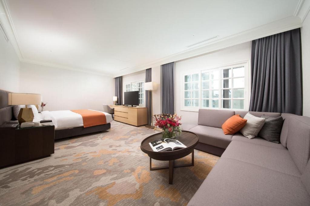 Spacious hotel room with a large bed, orange accents, a flat-screen TV on a wooden dresser, a sofa with cushions, a round coffee table with flowers and magazines, and large windows with gray curtains.