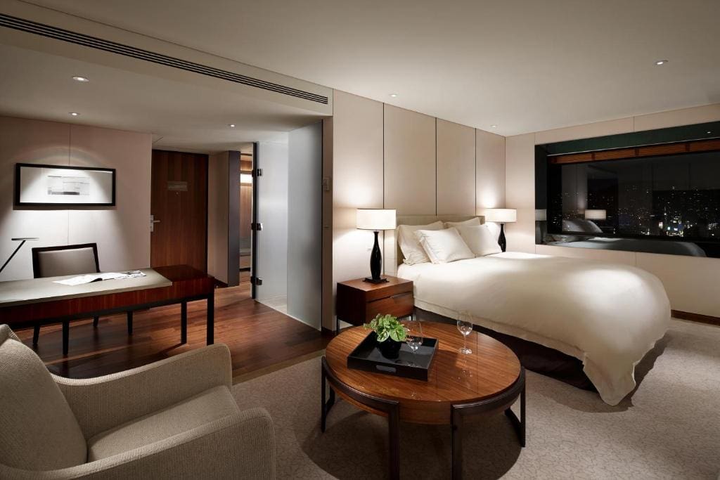 A modern hotel room with a large bed, a desk and chair, a round coffee table with a plant and glass, an armchair, bedside tables with lamps, and a large window showing a city night view.