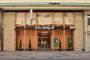 The entrance of Hotel Dormy Inn Annex, featuring stone and wood paneling, green potted trees, glass doors, and animal carvings above the doorway on a city street.
