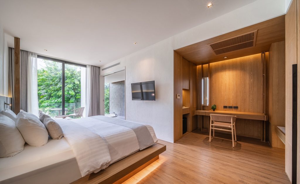 A modern hotel room with a large bed, white linens, wooden floors, a wall-mounted TV, a built-in wooden desk with a chair, and floor-to-ceiling windows overlooking greenery.