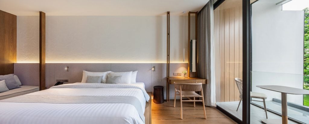 Modern hotel room with a large bed, white linens, a wooden desk and chair, sliding glass doors leading to a balcony with outdoor seating, and natural light filling the space.