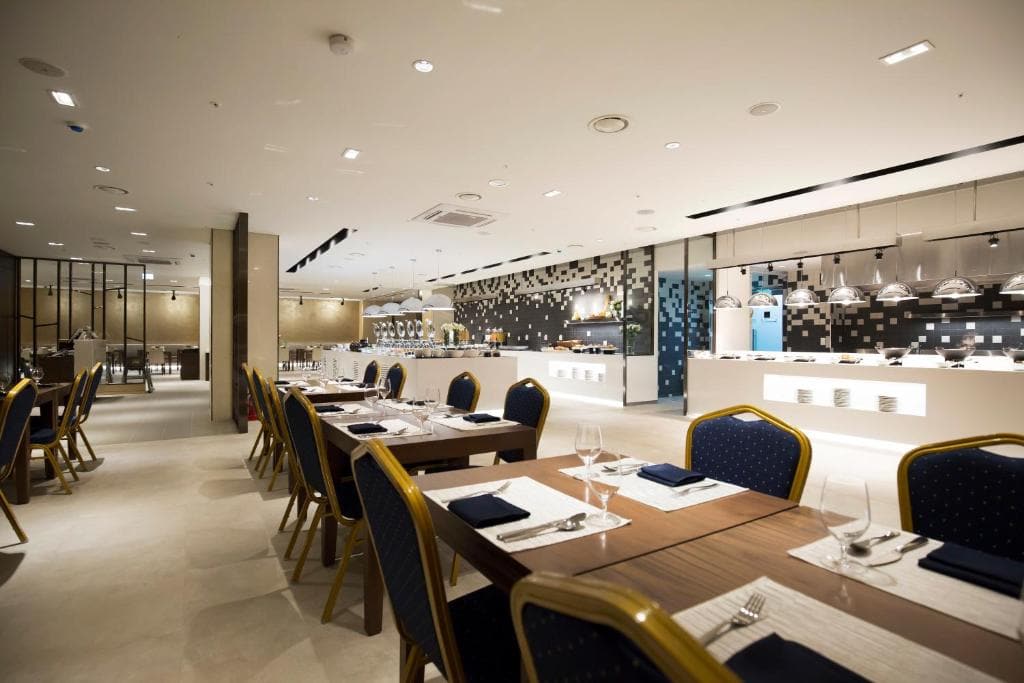 Modern, spacious restaurant with neatly set wooden tables and blue cushioned chairs. The buffet area features a long counter, food displays, and a stylish tiled wall in the background under bright lighting.