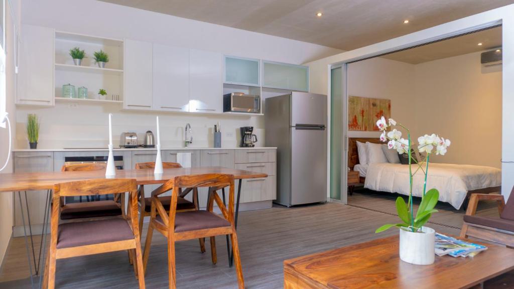 Modern open-plan apartment with a kitchen, wooden dining table and chairs, visible stainless steel appliances, and a bedroom separated by sliding glass doors. A white orchid sits on the coffee table in the foreground.