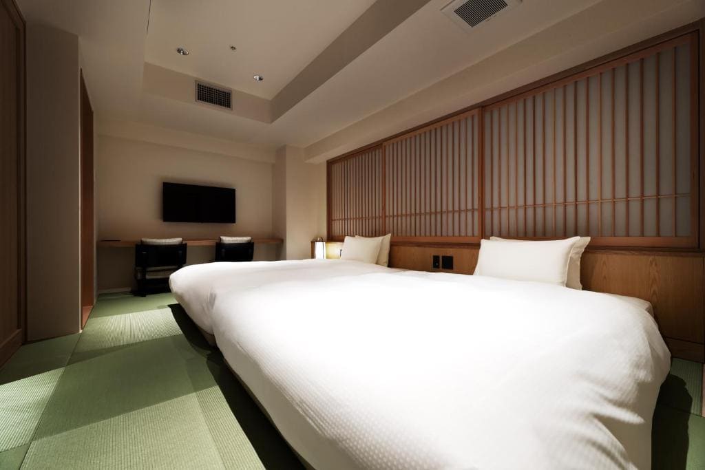 A modern hotel room with two large beds, a green tatami mat floor, wooden sliding panel doors, a flat-screen TV, and a desk with two chairs along the back wall. Warm lighting creates a cozy atmosphere.