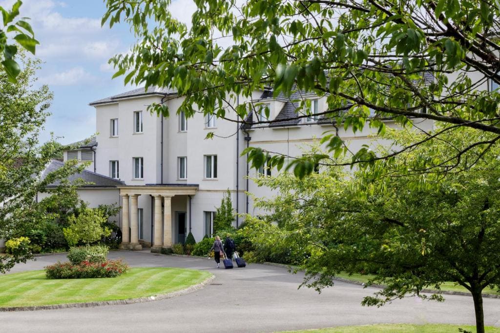 A modern three-story white hotel is surrounded by green lawns, trees, and a circular driveway. Two people with suitcases walk toward the entrance under a columned porch.