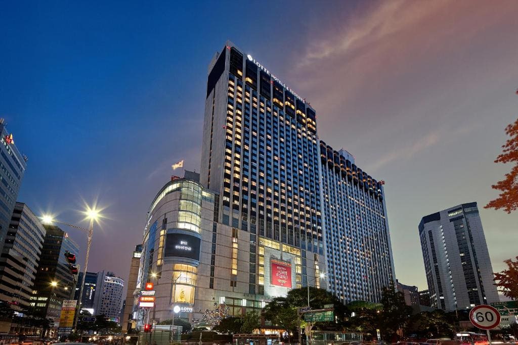 Tall, modern hotel building with many windows, illuminated at dusk in a cityscape with other high-rise buildings, streetlights, and a 60 km/h road sign in the foreground.