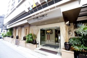 Street-level view of the entrance to The Key Bangkok hotel, with potted plants, glass doors, tall flagpoles above the canopy, and a clean, modern facade along a city sidewalk.