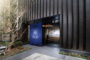 Entrance to MIMARU hotel with a dark facade, blue curtain displaying the MIMARU logo, and a glimpse of a mural and seating area visible through the doorway. Plants and trees frame the entrance.