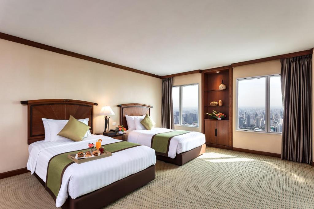 A hotel room with two single beds, each neatly made with white linens and green runners. There are decorative pillows, a tray with breakfast, large windows showing a city view, beige walls, and brown curtains.