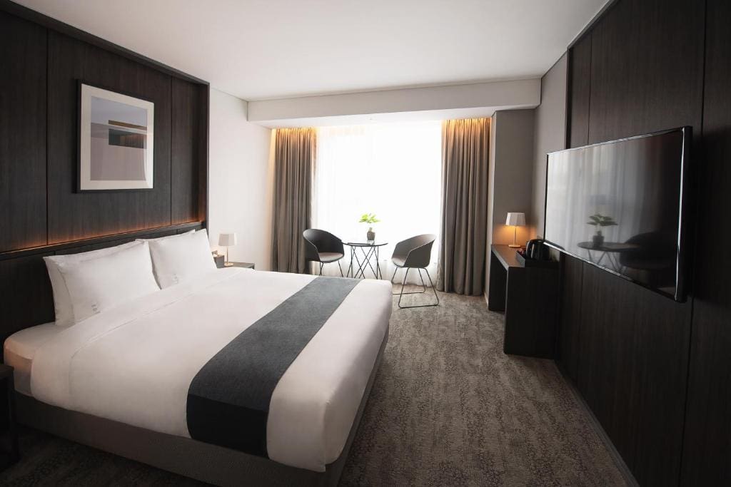Modern hotel room with a large bed, dark wood paneling, a wall-mounted TV, a small desk with a lamp, and a sitting area by a window with two chairs and a round table. Light streams in through sheer curtains.