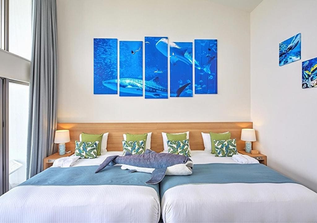 A bright hotel room with two beds, blue and white bedding, green accent pillows, a plush shark toy, and ocean-themed artwork featuring sharks on the wall above the beds.