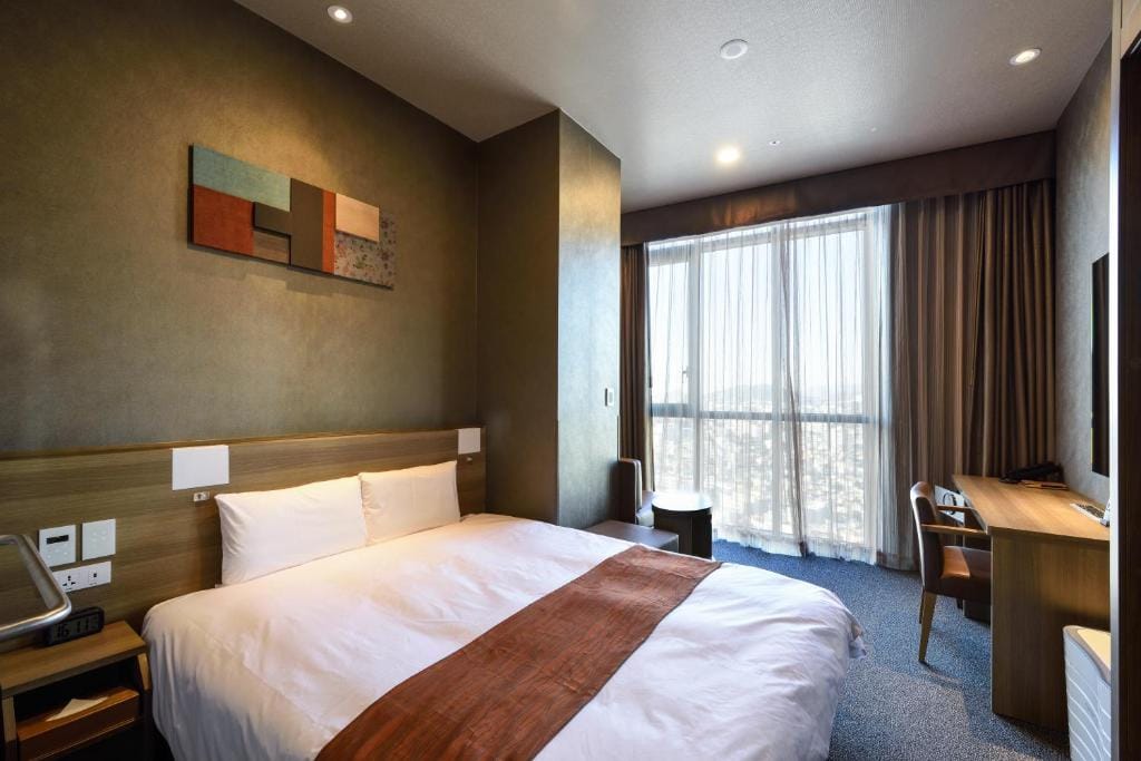 A modern hotel room with a double bed, brown accent runner, abstract wall art, desk, chair, and large window with sheer curtains letting in natural light.