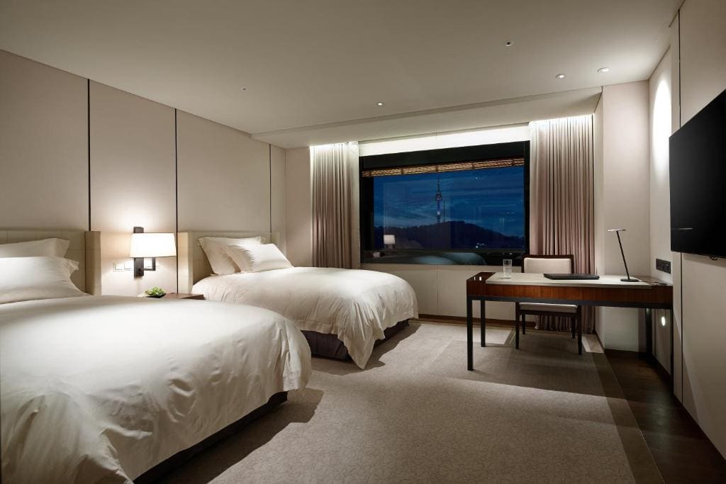 Modern hotel room with two double beds, a desk with a chair and lamp, large window with city view at dusk, neutral-toned walls and carpet, and a flat-screen TV mounted on the wall.