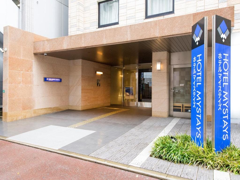 Entrance of HOTEL MYSTAYS with glass doors, beige stone walls, a covered walkway, signage in English and Japanese, and a small landscaped area with green plants in front.