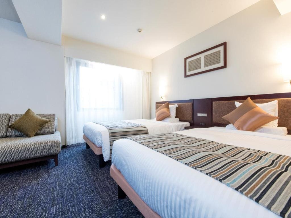A bright hotel room with two twin beds, each with striped runners and accent pillows, a patterned sofa with cushions, and a large window letting in natural light.